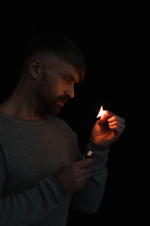 man with matchbox looking at lit match during energy blackout isolated on blackの写真素材
