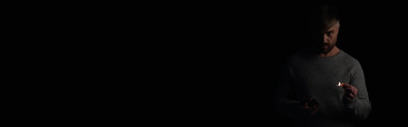 displeased man with matchbox and burning match looking at camera during electricity shutdown isolated on black, bannerの写真素材