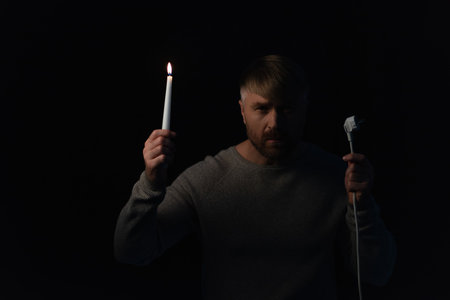 man with plug and burning candle looking at camera during electricity shutdown isolated on blackの写真素材