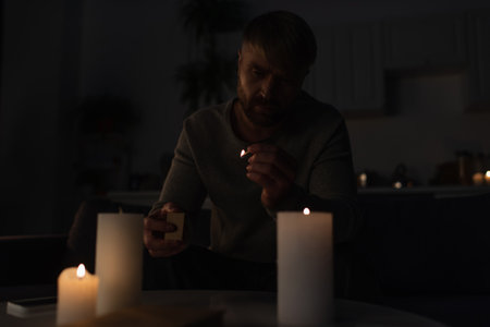 man holding lit match near burning candles while sitting in kitchen during power outageの写真素材
