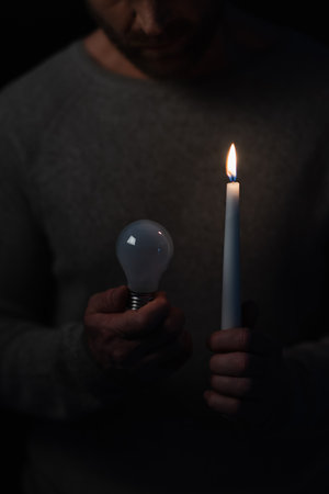 cropped view of man in darkness holding lit candle and electric bulb isolated on blackの写真素材