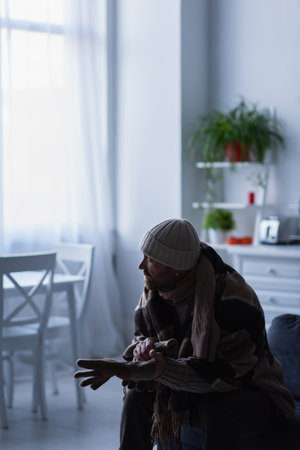 frozen man in knitted hat wearing warm gloves while sitting under blanket and looking away in twilightの写真素材