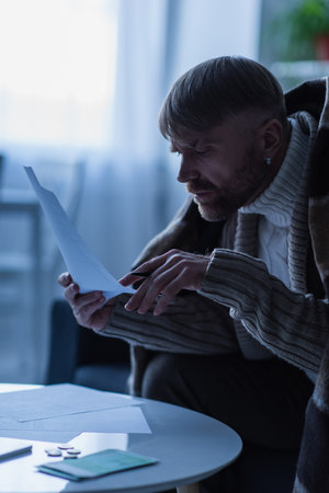 frozen man in warm sweater and blanket looking at payment bills during electricity shutdownの写真素材