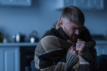 frozen man with closed eyes sitting in warm blanket during energy blackout at homeの写真素材