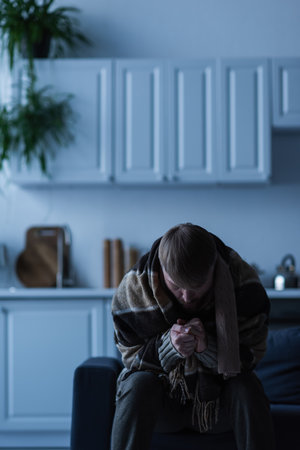 man wrapped in blanket warming hands while sitting in kitchen during power shutdownの写真素材