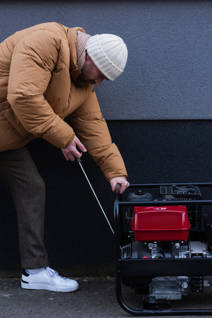 side view of man in down jacket and knitted hat starting electricity generator outdoors