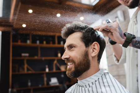 bearded brunette man smiling near hairdresser applying hair sprayの写真素材