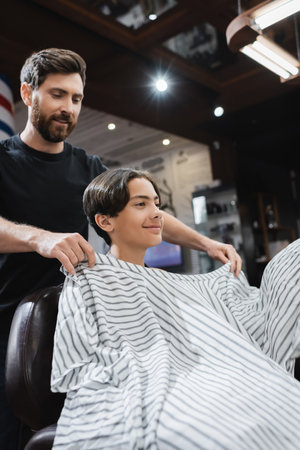 bearded hairstylist putting hairdressing cape on smiling teenager in barbershopの写真素材