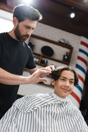 barber cutting hair of brunette teenage sitting in hairdressing capeの写真素材
