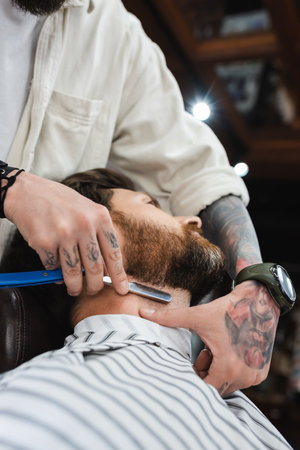 tattooed hairdresser shaving neck of bearded man with straight razorの写真素材