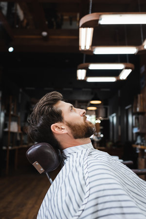side view of bearded brunette man sitting in hairdressing cape in barbershopの写真素材
