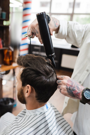 tattooed hairdresser in wristwatch drying hair of client in barbershop