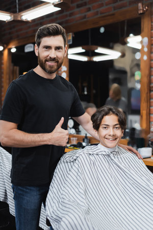 Positive barber showing like gesture and looking at camera near teenager in hairdressing capeの写真素材