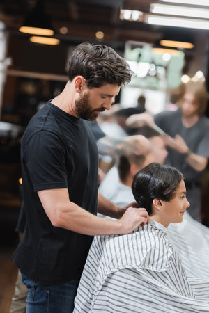 Side view of bearded hairdresser wearing cape on teen client in beauty salon