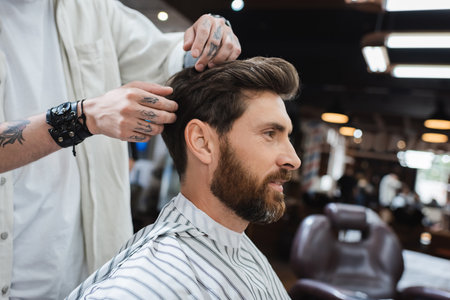 brunette bearded man in hairdressing cape near tattooed barber touching his hair in beauty salonの写真素材