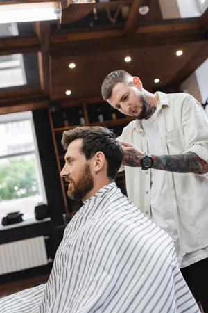 Smiling man in hairdressing cape sitting near blurred barber in barbershop