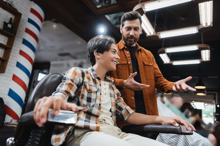 Low angle view of smiling teenage client sitting in armchair while barber talking in barbershop