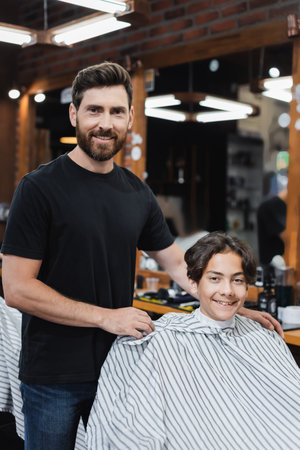 Smiling barber and teenage boy looking at camera in barbershopの写真素材