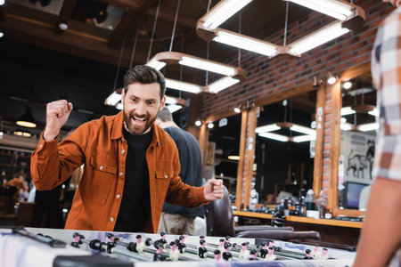 Excited man looking at table football near blurred son in barbershop