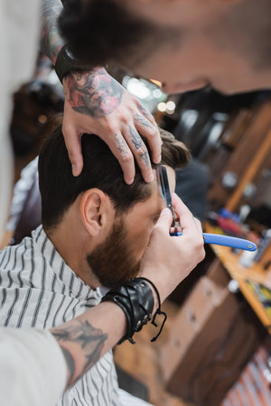 Tattooed barber trimming hair of client with straight razor in barbershopの写真素材