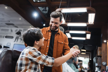 Smiling bearded barber holding hand of teenage client in barbershopの写真素材
