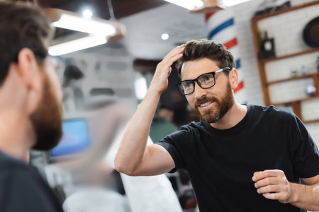 Smiling client in eyeglasses looking at mirror and touching hair in barbershop