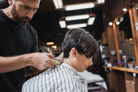 brunette teenage boy in hairdressing cape near bearded barber trimming his neckの写真素材