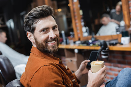 bearded man with glass of cappuccino and blurred cellphone smiling at camera in barbershopの写真素材