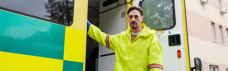 Paramedic standing near ambulance car and looking at camera outdoors, banner