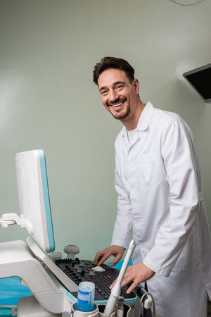 happy doctor in white coat looking at camera while operating ultrasound machine in clinicの写真素材