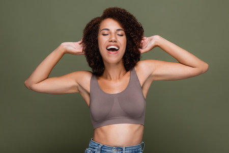 Happy african american woman in top touching curly hair isolated on greenの写真素材