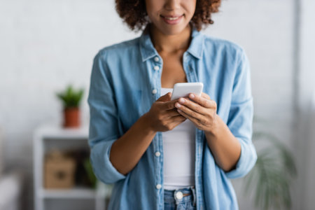 cropped view of curly african american woman smiling while messaging on smartphoneの写真素材