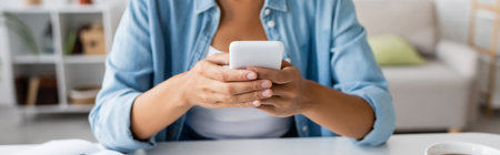 Cropped view of african american woman in casual clothes using smartphone near cup of tea at home, bannerの写真素材
