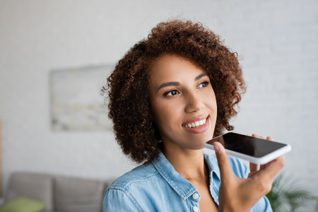 cheerful african american woman with curly hair recording voice message on smartphone