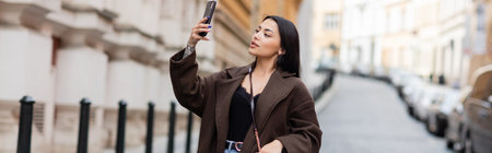 pretty brunette woman taking photo on cellphone on blurred street in prague, banner