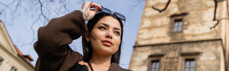 Low angle view of stylish brunette woman holding sunglasses on street in Prague, banner
