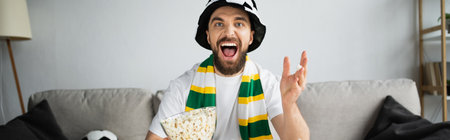 excited man in fan hat and scarf watching championship while holding bowl with popcorn, banner