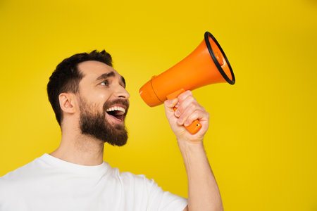 brunette bearded man shouting in megaphone while looking away isolated on yellowの写真素材