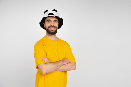 happy bearded man in football fan hat and yellow t-shirt standing with crossed arms isolated on grey