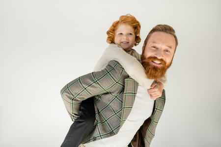 Smiling redhead kid piggybacking on bearded dad isolated on greyの写真素材