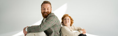 Cheerful red haired man in warm jumper looking at bearded parent on grey background with light,...