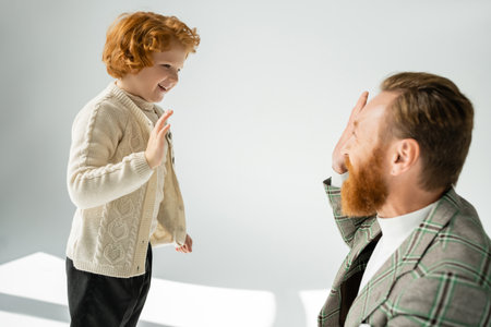 Smiling red haired kid in knitted jumper giving high five to father on grey backgroundの写真素材