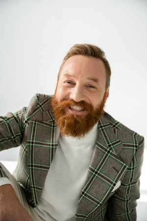 Positive bearded man in plaid jacket looking at camera on grey background