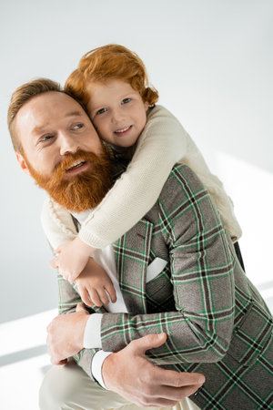 Smiling redhead boy hugging stylish parent on grey background with sunlightの写真素材