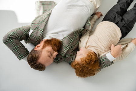 top view of redhead father and son in trendy outfit looking at each other while lying on grey...