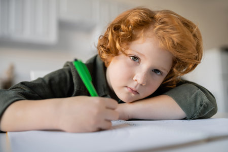bored redhead boy with pen looking at camera near blurred notebookの写真素材