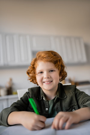 happy redhead kid with pen looking at camera while doing homework on blurred foregroundの写真素材