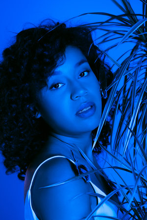 curly african american woman with butterflies on body looking at camera near palm leaves isolated on...