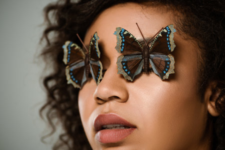 close up of curly african american woman with butterflies on eyes isolated on greyの写真素材
