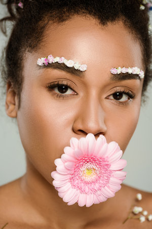 close up of african american woman with flowers on eyebrows and hair with gerbera in mouth isolated...